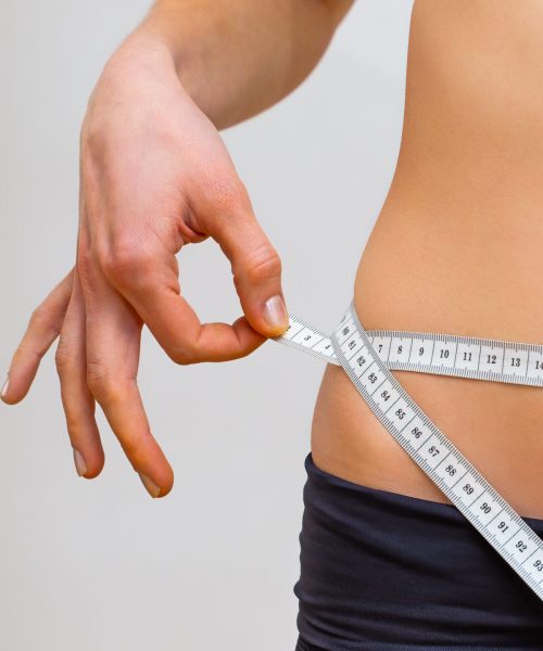 Young woman measuring her waistline in a gym with a tape. Concept of weight loss and fat removing. Slim abdomen of a young adult after training from close-up.